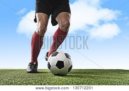 close up legs and feet of football player in red socks and black shoes running and dribbling with the ball playing outdoor on green grass pitch under a blue sky