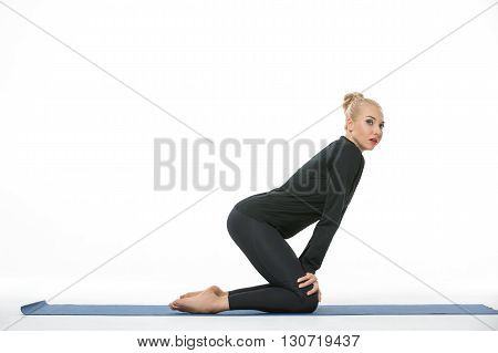 Sexy blonde girl in the sportswear stands on the knees on a blue gymnastic mat on the white background in the studio. She wears black pants and black long sleeve t-shirt. She is barefoot. Her hands are on the knees. She tilts her body forward. She looks t
