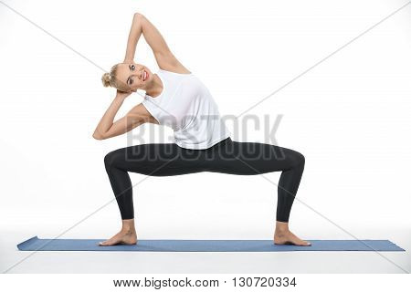 Smiling blonde girl in the sportswear squats with divorced feet on a blue gymnastic mat on the white background in the studio. She wears black pants and white sleeveless t-shirt. She is barefoot. She holds her hands on the nape and tilts torso to the righ