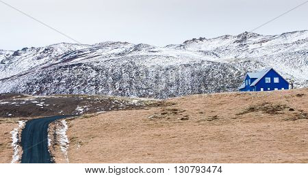 Typical Icelandic landscape at Reykjanes Peninsula with mountains covered in snow and empty asphalt road leading to a blue cottage house