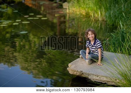 8-9 years boy sitting cross-legged on the shore of a picturesque pond. He smiled looking at the camera. The water reflects the sky and reeds.