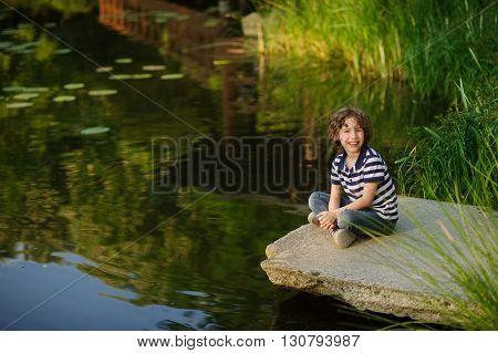 On the shore of the pond is sitting cross-legged boy. He smiles. The picturesque pond surrounded by green bulrush