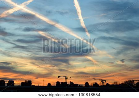 Vapor trails and thin clouds in a blue and orange sky with a rooftop skyline at the bottom