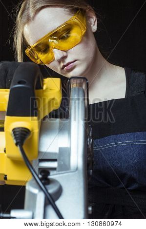 young beautiful woman in overalls and glasses with disk saw preparing for cutting. Photo on black background. close-up portrait.
