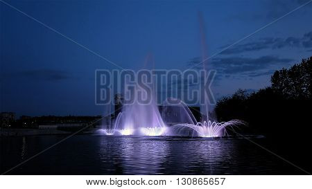 Fountain with bright illumination on the water pond or river with beautiful reflection at the evening or night. Beauty and calm tranquility urban recreational concept.