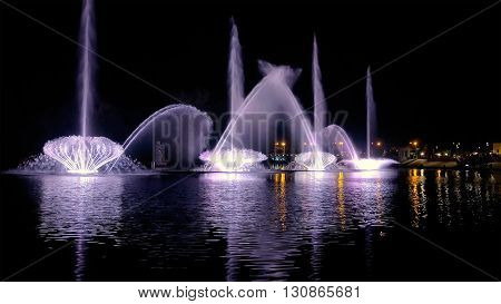 Fountain with bright illumination on the water pond or river with beautiful reflection at the evening or night. Beauty and calm tranquility urban recreational concept.