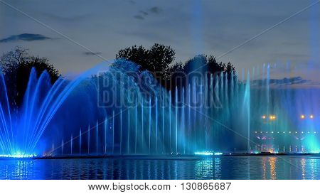 Amazing fantastic colorful fountain with bright illumination on the water pond or river with beautiful reflection at the evening or night. Beauty urban recreational concept.