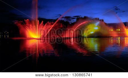 Amazing fantastic colorful fountain with bright illumination on the water pond or river with beautiful reflection at the evening or night. Beauty urban recreational concept.