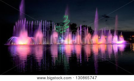 Amazing fantastic colorful fountain with bright illumination on the water pond or river with beautiful reflection at the evening or night. Beauty urban recreational concept.
