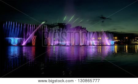 Amazing fantastic colorful fountain with bright illumination on the water pond or river with beautiful reflection at the evening or night. Beauty urban recreational concept.