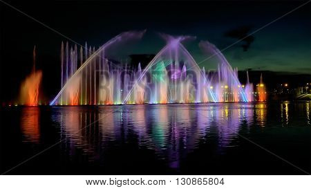Amazing fantastic colorful fountain with bright illumination on the water pond or river with beautiful reflection at the evening or night. Beauty urban recreational concept.