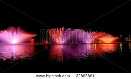 Amazing fantastic colorful fountain with bright illumination on the water pond or river with beautiful reflection at the evening or night. Beauty urban recreational concept.