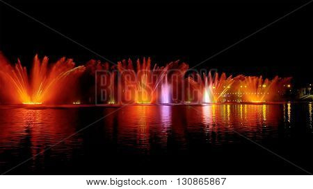 Amazing fantastic colorful fountain with bright illumination on the water pond or river with beautiful reflection at the evening or night. Beauty urban recreational concept.