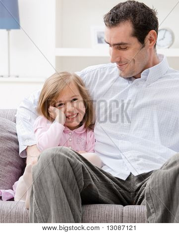 Bambina indossa abito rosa seduto sul divano abbracciato dal padre, sorridente.