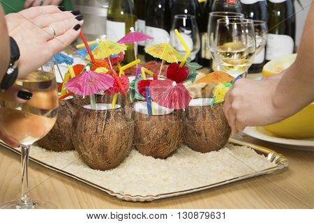 Coconut cocktail with colorful straws and colored paper palm trees wine bottles and woman's hands with wineglasses