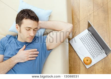 Mladý muž spí na pohovce doma, notebook na podlaze.  Pohled na stropní.
