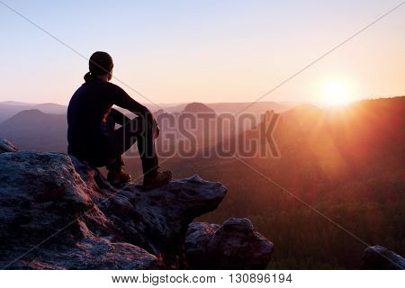 Tired Adult Hiker In Black Trousers, Jacket And Dark Cap Sit On Cliff Edge And Looking To Colorful M
