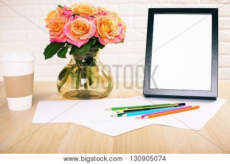 Roses in vase blank picture frame paper sheets with colorful pencils and take away coffee cup on wooden desktop and white brick wall background. Mock up