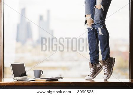 Woman's legs in grey sneakers standing on windowsill with blank laptop screen coffee cup and notepads on blurry city background. Mock up