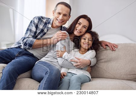 Happy Together. Beautiful Content Curly-haired Girl Smiling And Sitting On The Couch With Her Parent