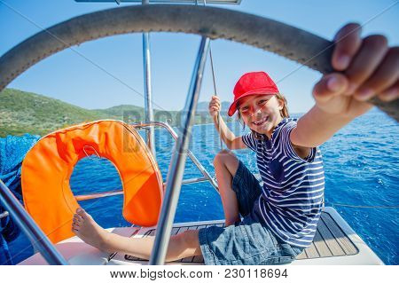 Cute Boy Captain On Board Of Sailing Yacht On Summer Cruise. Travel Adventure, Yachting With Child O