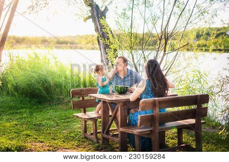 Family In Summer On A Picnic Near A Pond, Water. Family Holiday In Nature. A Little Daughter Feeds H