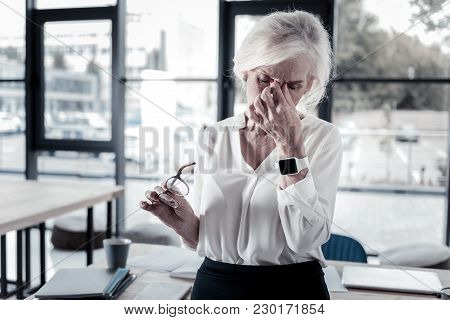 I Am Tired. Serious Blonde Woman Bowing Head And Touching Bridge Of Nose While Keeping Eyes Closed