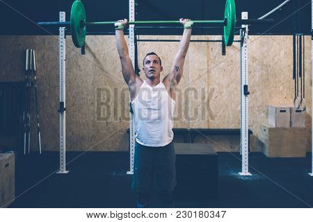 Young Athlete Man Doing Thruster Workout With The Barbell Over The Head At The Box Gym