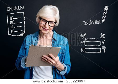 Check List. Calm Clever Attentive Woman Wearing Glasses And Standing With A Convenient Clipboard In 