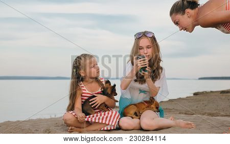 Two Teenage Are Sitting On A Sandy Beach. Near Them Is The Mother Of The Girls. The Sisters Show The