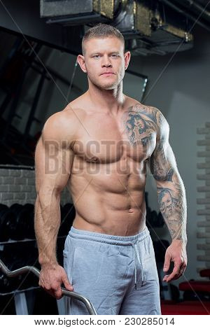 Muscular Shirtless Man With Blue Eyes And Tattoo Poses With Ez Curl Bar In A Gray Pants In A Gym