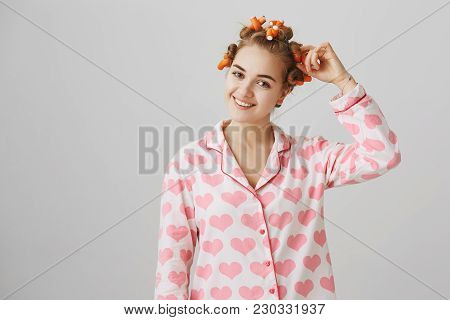 Indoor Shot Of Positive Cosy Woman Wearing Curlers And Lovely Pyjamas With Heart Print, Touching Wav