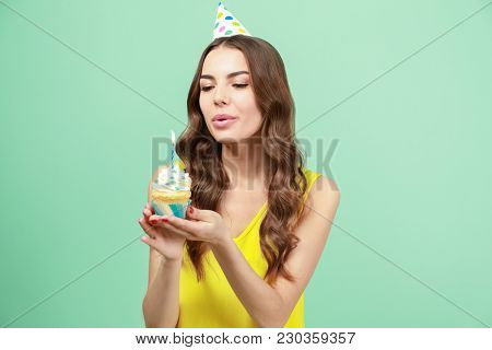 Beautiful young woman snuffing out candle on birthday cupcake against color background