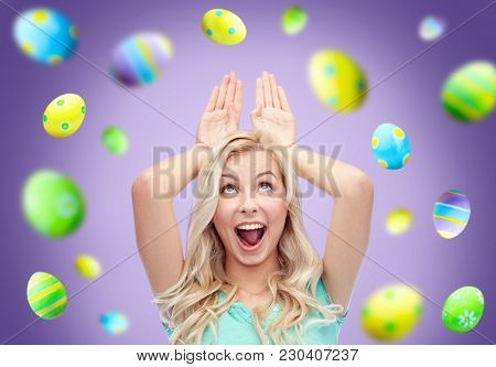 easter, holidays and people concept - happy smiling young woman making bunny ears over ultra violet background with colored eggs