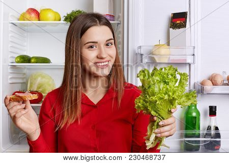 Attractive Woman Wears Fashionable Red Blouse, Stands Near Opened Fridge, Holds Lettuce And Sandwhic