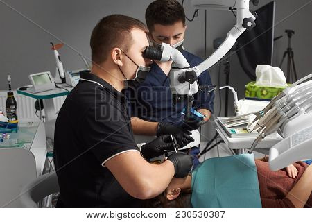 Male Dentist With An Assistant In A Black Mask And Gloves Treats A Patient Using A Microscope. Moder