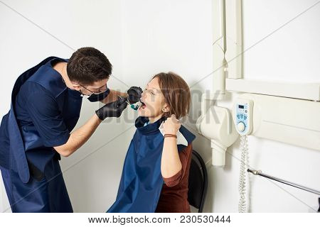 Dentist In A Blue Uniform Is Curing The Patient Making An X-ray Shot In A White Dental Office