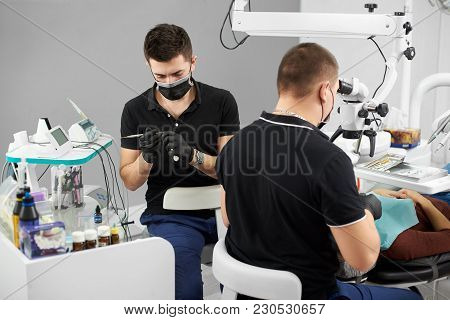 Male Dentist In A Black Uniform Is Examining The Patient In A Dental Chair While Another Male Dentis