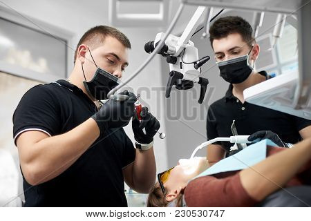 Stomatologist Prepares A Tool To Treat A Patient, A Young Assistant Helps Him. Both Are Dressed In B