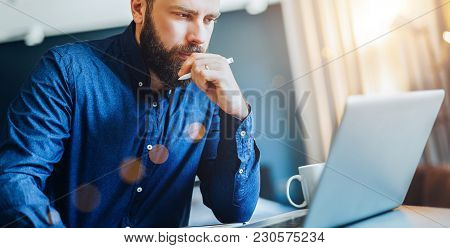 Young Serious Bearded Businessman Sitting At Table In Front Of Computer, Looking At Screen, Holding 
