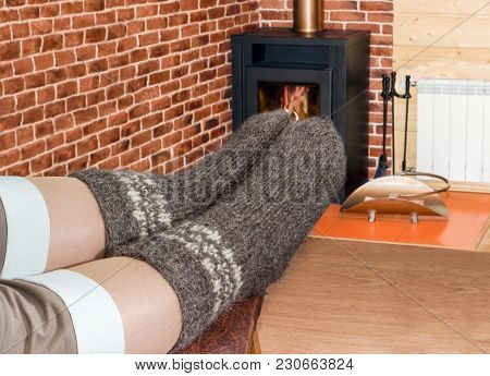 Pair Of The Thick Gray Wool Hand-knitted Socks On Women Legs On The Background Of A Burning Fireplac