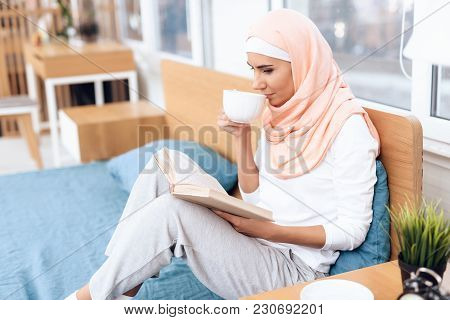 An Arabian Woman Is Drinking Tea And Reading A Book While Sitting On The Bed. She Rests At Home.