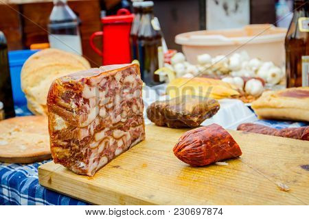 Headcheese And Smoked Sausage On A Wooden Chopping Board At Outdoor Kitchen, People In Background Ar