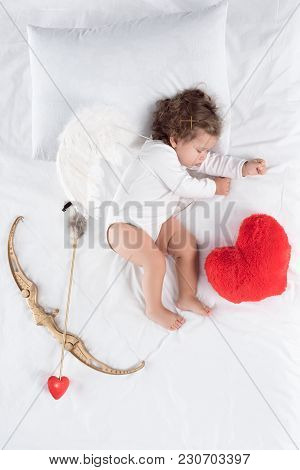 Little Baby With Wings Lying On Bed With Heart Pillow, Bow And Arrow