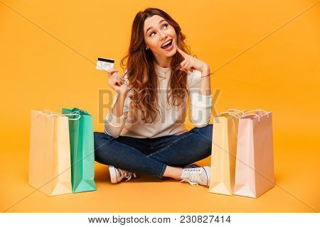 Pensive intrigued brunette woman in sweater sitting on the floor with packages while holding credit card and looking up over yellow background