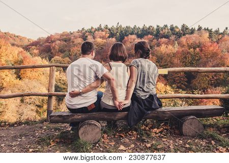 Three Young Beautiful People Sit On A Wooden Bench And Enjoy The Autumn Landscape, The Concept Of A 