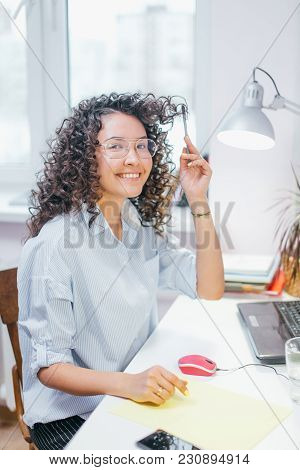 Pozitive Girl With Glasses And Dark Wavy Hair Touching Them With Pen Indoors. Perspective Plan