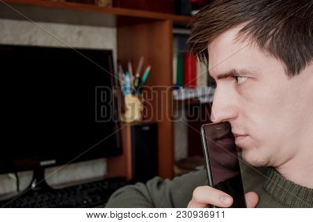 A Thoughtful Guy With A Smartphone At His Mouth, Sits At His Desk, On A Black Monitor Background