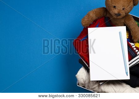 Donation Box With Clothing, Books, Children Toys With Blank Notebook Page And Pencil On Blue Backgro
