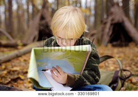 Little Boy Scout Is Orienteering In Forest. Child Is Sitting On Fallen Tree And Looking On Map On Ba